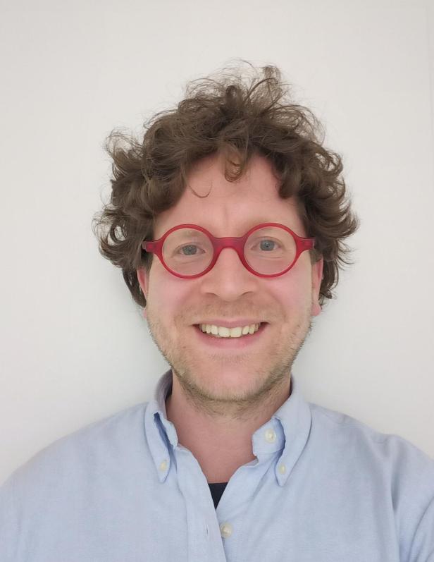 Mann mit lockigen braunen Haaren, roter Brille und hellblauem Hemd lächelt vor weißem Hintergrund.