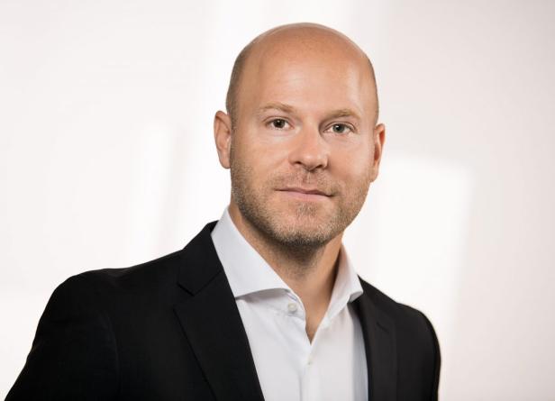 Ein Mann mit Glatze und kurzem Bart trägt ein weißes Hemd und einen dunklen Anzug vor hellem Hintergrund.