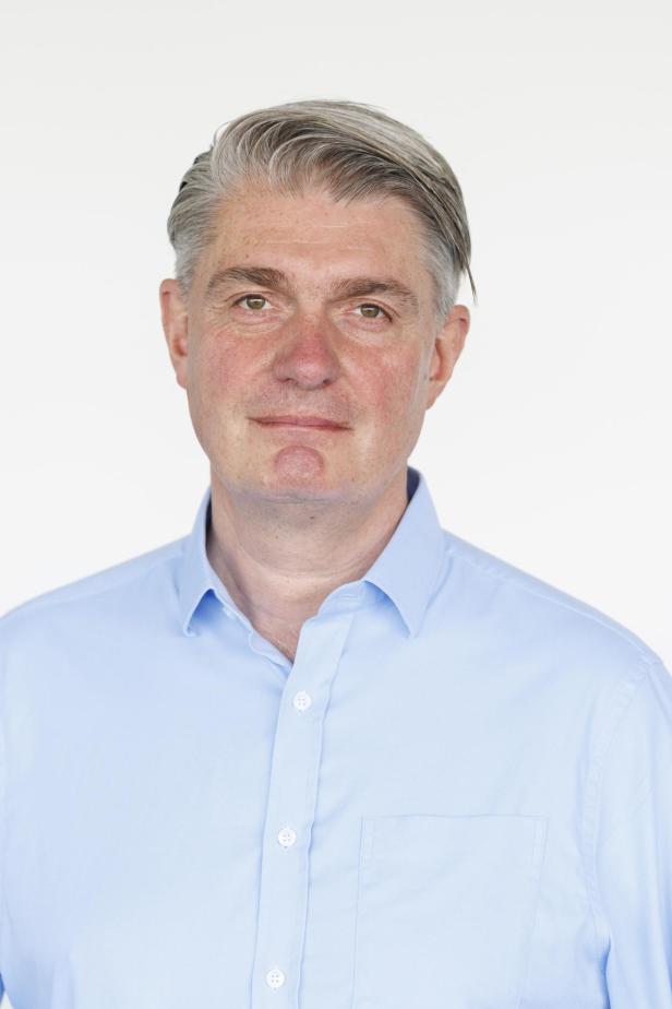 Mann mit grauem Haar und hellblauem Hemd blickt neutral nach vorne vor hellem Hintergrund.