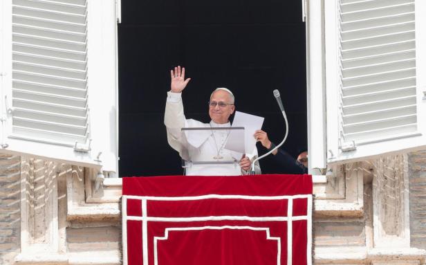 Papst Leo XIV. winkt von einem Fenster mit roten Vorhängen.