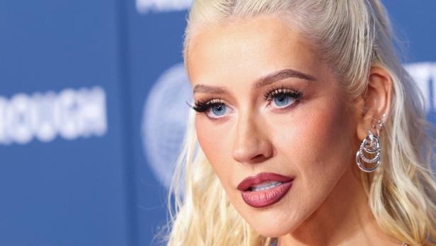 Christina Aguilera mit blonden Haaren, blauem Lidschatten und Ohrringen vor blauem Hintergrund.