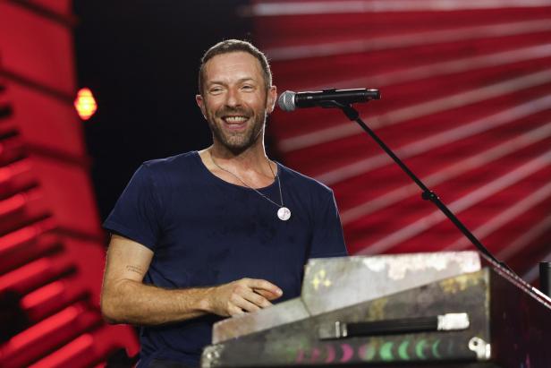 Chris Martin lächelt am Keyboard vor einem Mikrofon auf der Bühne.