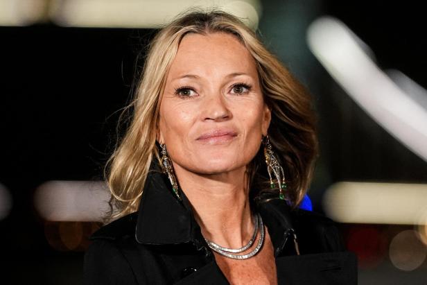 Kate Moss mit blonden Haaren, Ohrringen und Halskette vor dunklem Hintergrund.