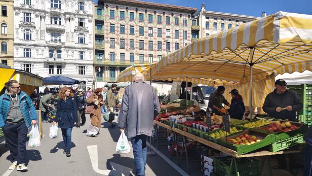 Menschen kaufen bei sonnigem Wetter an Marktständen mit Obst und Gemüse vor historischen Gebäuden ein.