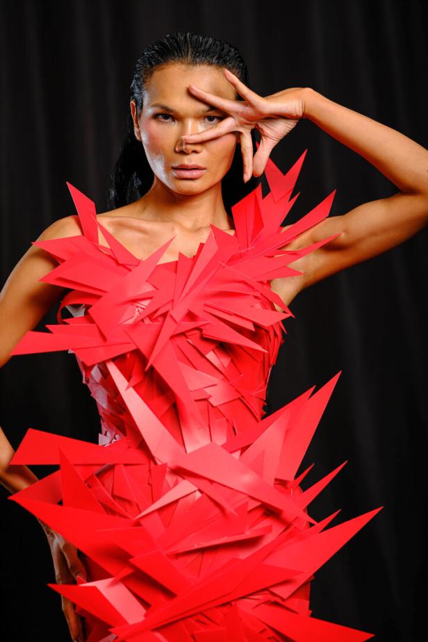 Eine Frau mit dunklen Haaren trägt ein rotes Kleid aus spitzen Formen und hält eine Hand vor ihr Gesicht.