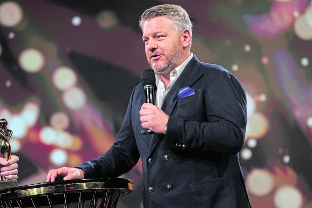 Ein Mann im Anzug spricht mit einem Mikrofon in der Hand an einem runden Tisch mit einer Trophäe.