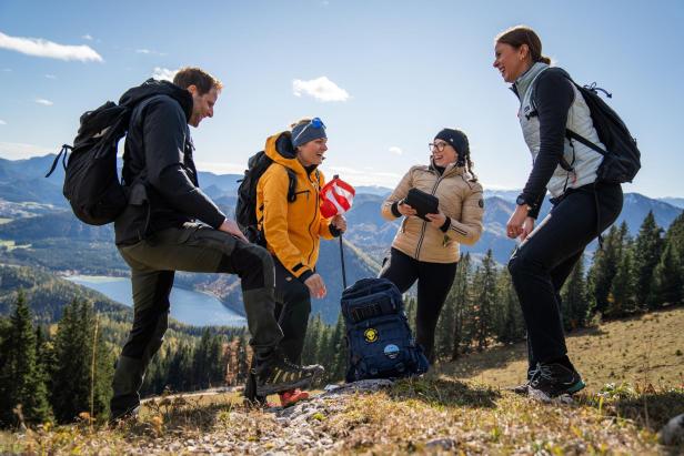Vier Personen mit Wanderausrüstung rätseln gemeinsam auf einem Berg mit Seeblick.