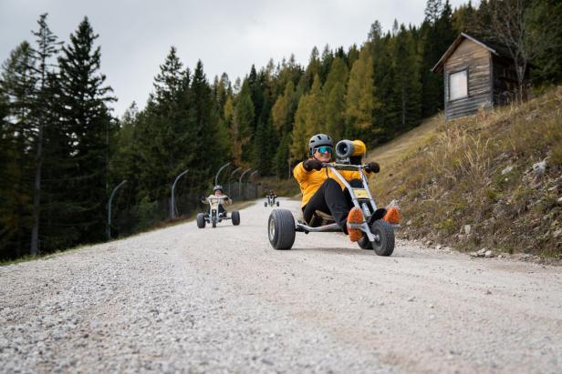 Drei Personen fahren mit Mountaincarts eine Schotterstraße durch bewaldetes Gelände hinab.