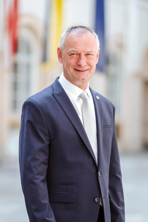 Ein älterer Mann im dunkelblauen Anzug mit weißer Krawatte steht draußen und lächelt in die Kamera.