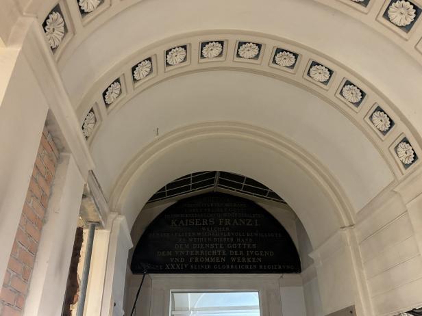 Gewölbte Decke mit floralen Verzierungen und einer schwarzen Gedenktafel mit goldener Inschrift über einem Durchgang.