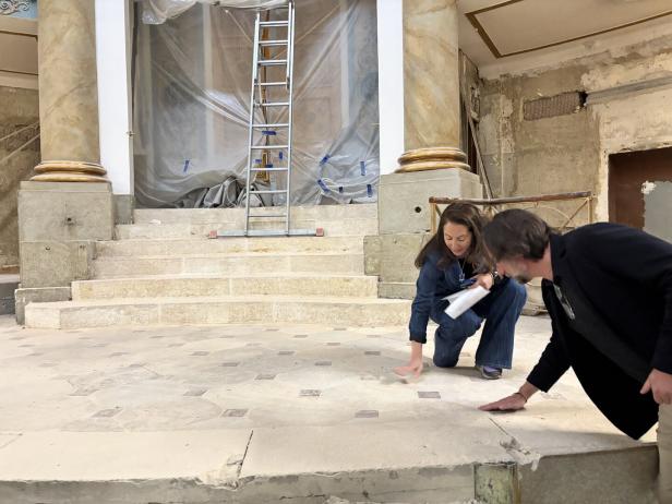 Zwei Personen begutachten kniend einen beschädigten Steinboden vor einer Treppe in einem renovierungsbedürftigen Gebäude.
