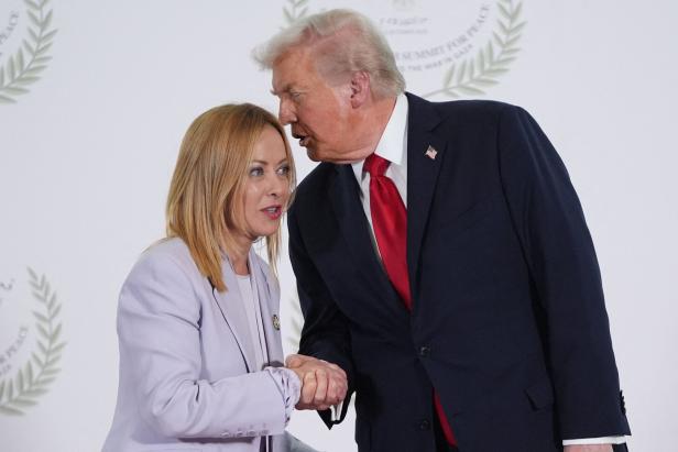 Donald Trump schüttelt Giorgia Meloni die Hand und beugt sich ihr zu, während sie sich vor einem hellen Hintergrund mit Lorbeerkranz-Symbolen ansehen.