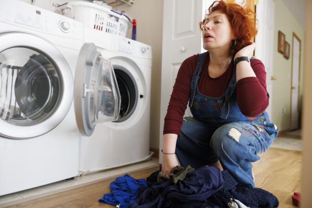 Eine erschöpfte Frau sortiert Wäsche vor einer Waschmaschine im Haushalt.
