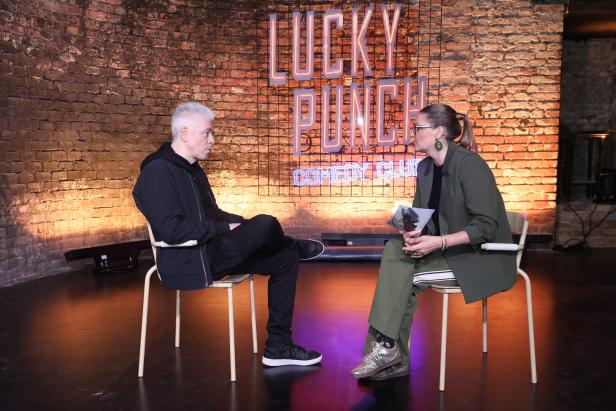 Zwei Personen sitzen sich auf einer Bühne gegenüber, im Hintergrund leuchtet ein „Lucky Punch Comedy Club“-Schriftzug.