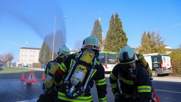 Drei Feuerwehrleute in Schutzkleidung und mit Atemgeräten löschen bei Sonnenschein einen Brand auf einer Straße.
