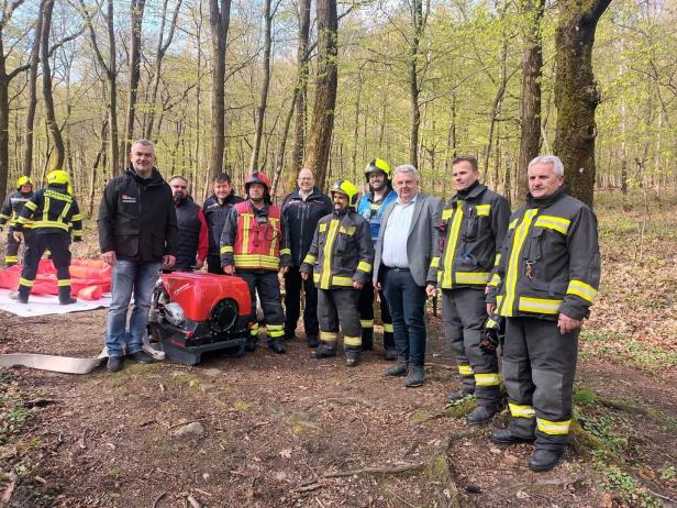 Mehrere Feuerwehrleute und weitere Personen stehen im Wald neben Ausrüstung und einem roten Gerät.