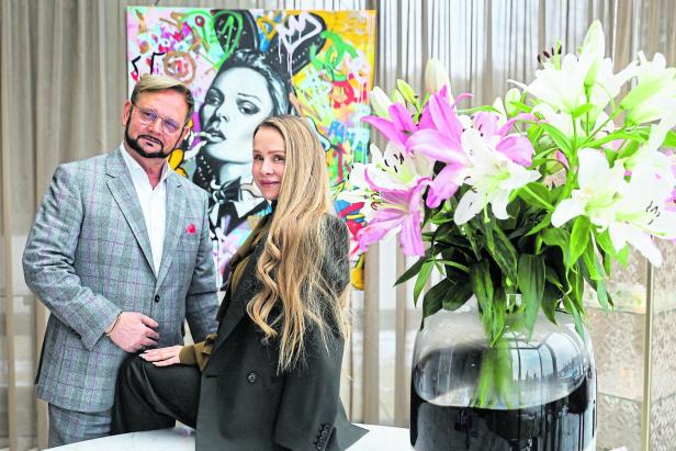 Ein Mann und eine Frau stehen vor einem bunten Gemälde, daneben steht eine große Vase mit weißen und rosa Lilien.