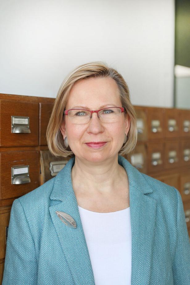 Eine Frau mit Brille und blauem Blazer steht vor einem alten Karteikasten.