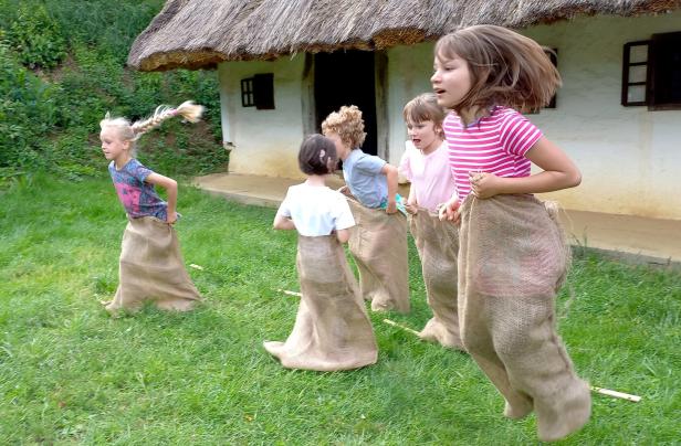 Fünf Kinder machen ein Sackhüpfen auf einer Wiese vor einem Haus mit Strohdach.