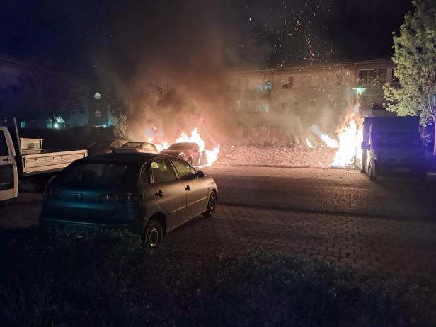 Mehrere Autos stehen auf einem Parkplatz, während zwei Fahrzeuge in Flammen stehen und dichter Rauch aufsteigt.