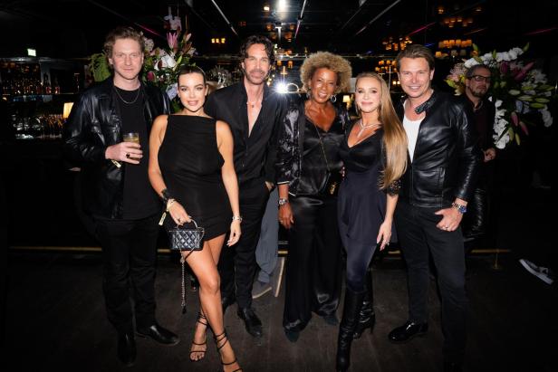 Sechs elegant gekleidete Personen posieren gemeinsam in einem Club vor Blumenarrangements.