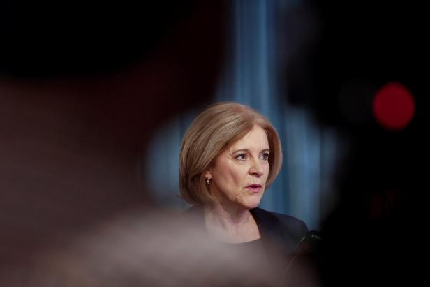 Eine Frau mit blonden Haaren spricht vor einem Mikrofon, im Hintergrund sind unscharfe Vorhänge zu sehen.