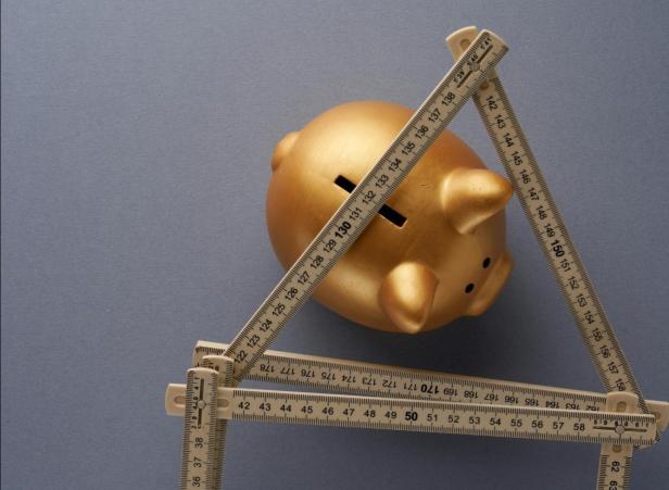 Ein goldenes Sparschwein liegt auf einer blauen Fläche und ist von einem aufgeklappten Zollstock umrahmt.