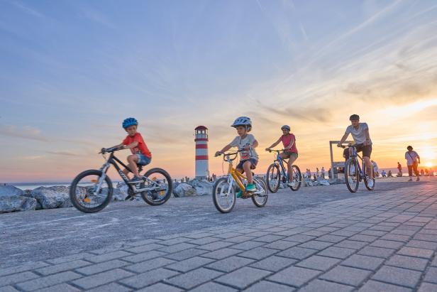 Vier Personen fahren bei Sonnenuntergang mit Fahrrädern an einem Leuchtturm am Wasser entlang.