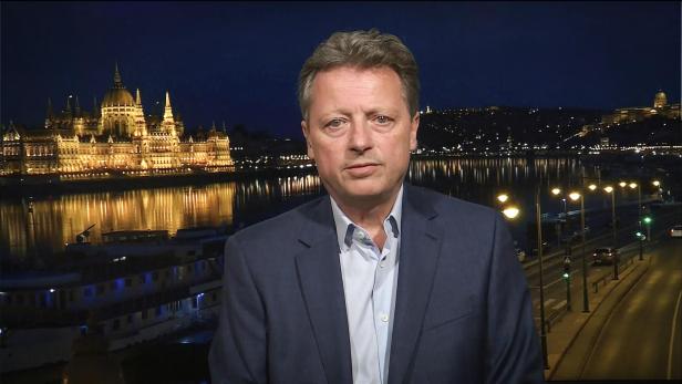 Ein Mann im Anzug steht vor einer nächtlich beleuchteten Stadt mit einem großen, angestrahlten Gebäude am Fluss.