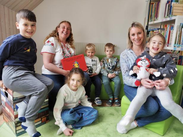 Mehrere Kinder und zwei Erwachsene sitzen lachend in einer gemütlichen Leseecke mit Büchern und einem Stofftier.