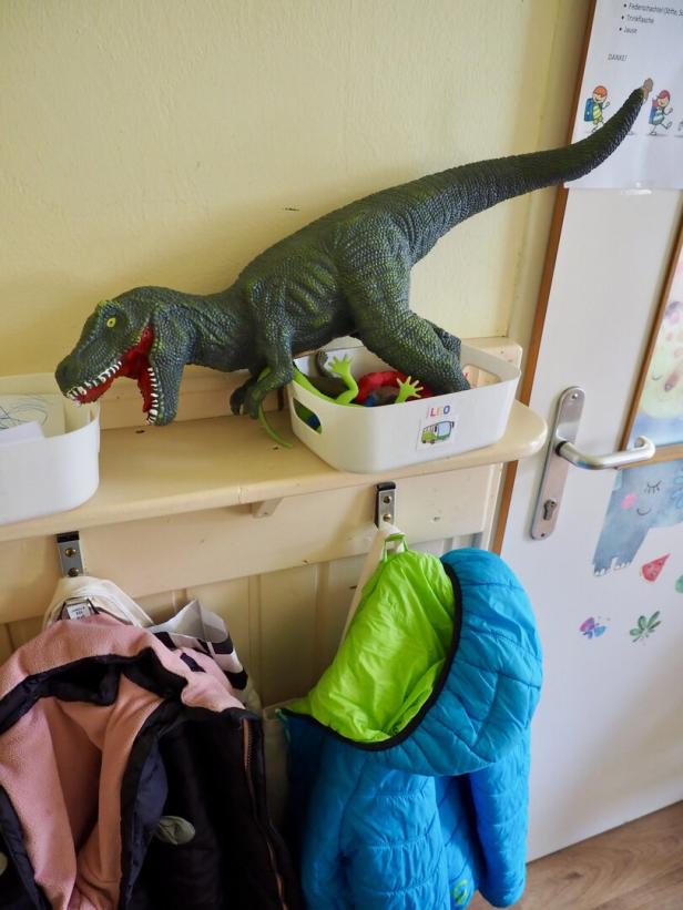 Ein großer Dinosaurier-Spielzeug steht auf einer Kiste mit weiteren Spielsachen über einer Garderobe mit Jacken.