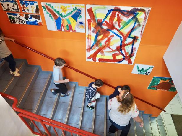 Mehrere Kinder gehen eine Treppe mit orange gestrichener Wand hinauf, an der bunte Kinderzeichnungen hängen.