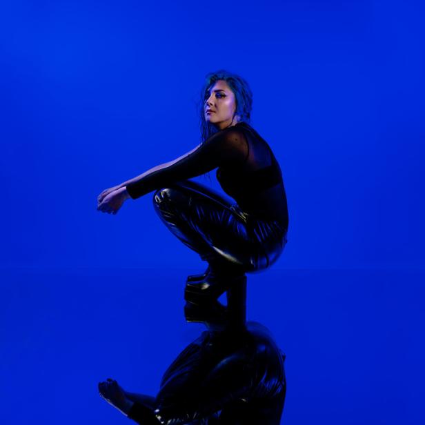 Paenda hockt in schwarzer Kleidung vor blauem Hintergrund.