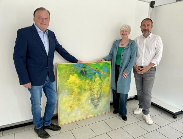 Drei Personen stehen nebeneinander und halten gemeinsam ein buntes abstraktes Gemälde mit überwiegend grünen und gelben Farben.
