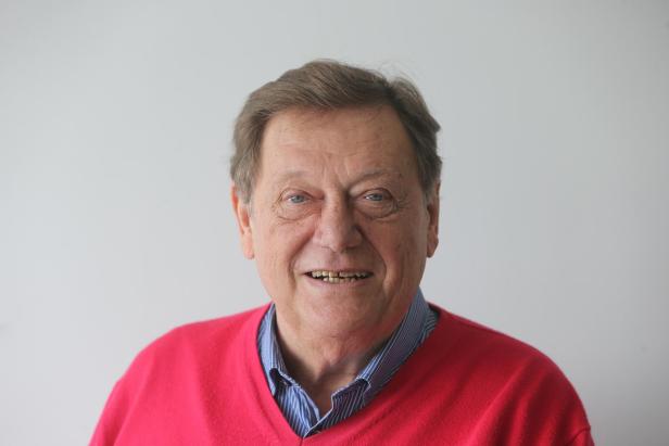 Älterer Mann mit kurzen braunen Haaren und rotem Pullover lächelt vor hellem Hintergrund.