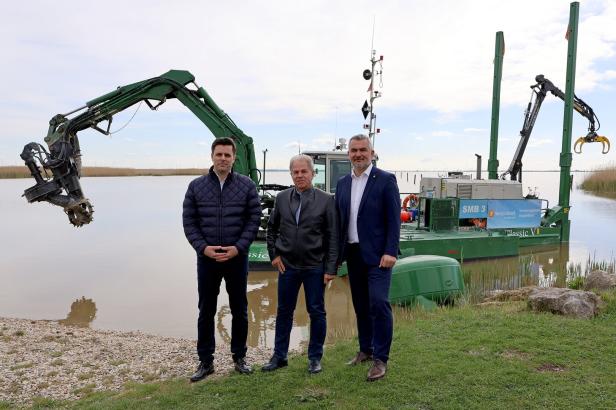 Drei Männer stehen am Ufer vor einem grünen Arbeitsboot mit zwei großen Greifarmen auf einem ruhigen See.