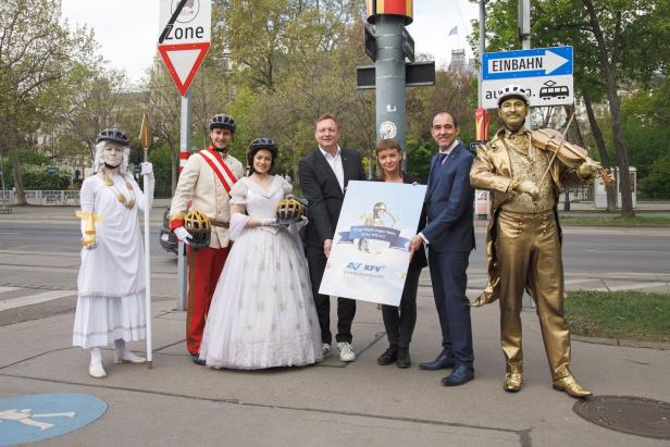 Eine Gruppe von Personen, darunter verkleidete Figuren und KFV-Vertreter, posiert mit einem Schild für eine Verkehrssicherheitskampagne.
