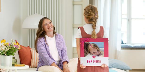 Ein Mädchen hält ein Fotobuch mit der Aufschrift „Für Mama“ hinter dem Rücken, Mutter lächelt.