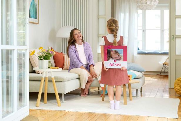 Ein Mädchen hält ein Fotobuch mit der Aufschrift „Für Mama“ hinter dem Rücken, Mutter lächelt.