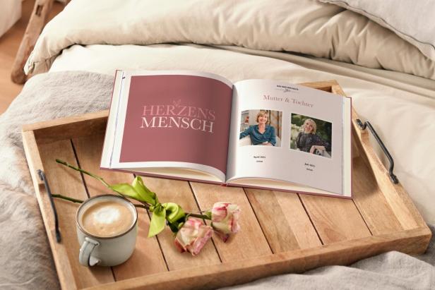 Auf einem Tablett liegen ein geöffnetes Fotobuch, eine Tasse Kaffee und eine Rose auf einem Bett.