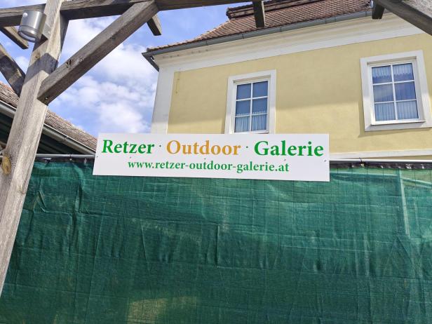 Schild der Retzer Outdoor Galerie vor gelbem Haus und grünem Bauzaun bei Sonnenschein.