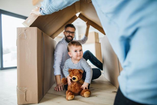 Ein kleines Kind mit einem Teddybär sitzt mit einem Erwachsenen in einem aus Kartons gebauten Haus.