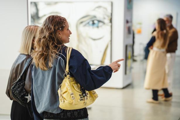 Zwei Frauen betrachten gemeinsam Kunstwerke in einer Galerie, eine zeigt auf ein Bild im Hintergrund.