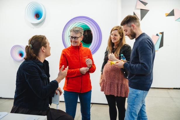 Vier Personen unterhalten sich in einer Galerie mit bunten, abstrakten Kunstwerken an den weißen Wänden.