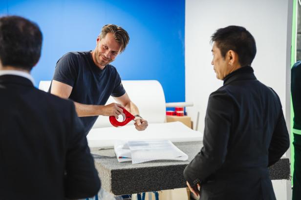 Ein Mann befestigt mit rotem Klebeband Luftpolsterfolie auf einem Tisch, während zwei weitere Personen zuschauen.