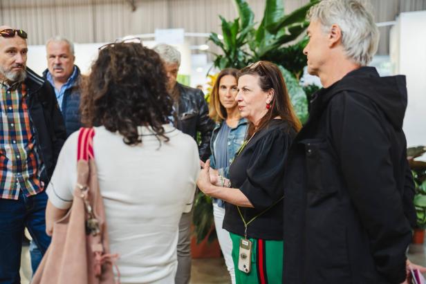 Eine Frau spricht in einer Gruppe von Erwachsenen, umgeben von großen grünen Pflanzen in einem Innenraum.