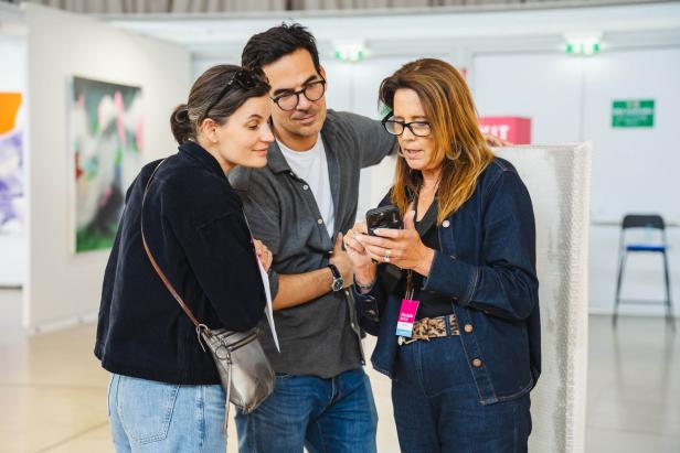 Drei Personen betrachten gemeinsam ein Smartphone in einer modernen, hellen Umgebung mit Kunstwerken an den Wänden.