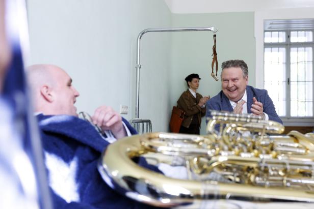 Michael Ludwig im Anzug applaudiert lachend einem anderen Mann, der im Bett liegt und ein Blasinstrument hält.