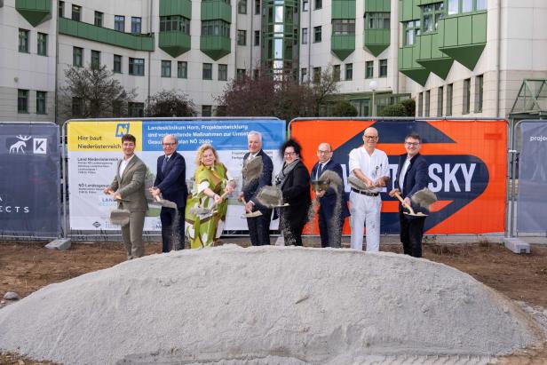 Sieben Personen stehen nebeneinander und schaufeln Sand vor einem Gebäude mit grünen Fassadenelementen.