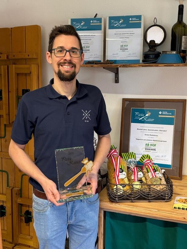 Ein Mann mit Brille hält eine Auszeichnung, im Hintergrund Urkunden und ein Korb mit prämierten Nudelprodukten.